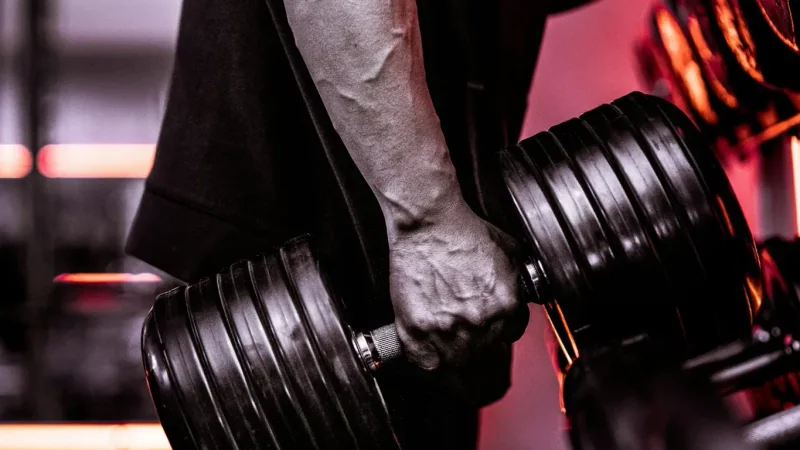 Forearm and hand gripping a heavy dumbbell in front of the dumbbell rack, red gym lighting