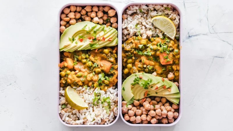 Meal-prep containers with rice, chickpeas, vegetables and avocado on a light stone surface.